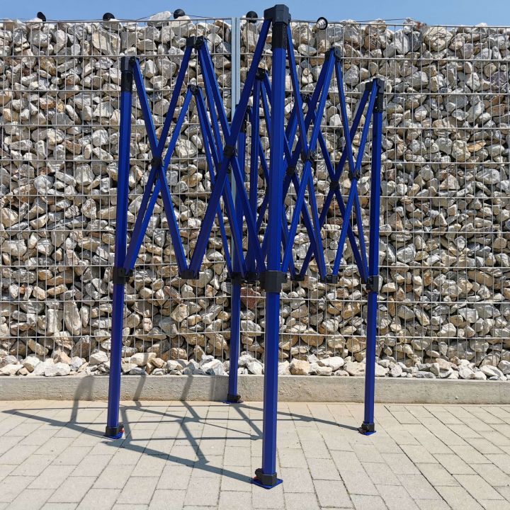 Αναδιπλωμένη, αλλά μη πυροβολημένη μπλε κατασκευή (σκελετός) σκηνής πάρτι τύπου ψαλίδι, που στέκεται σε πλακόστρωτο με βότσαλα στο βάθος (δίπλα στον φράχτη).